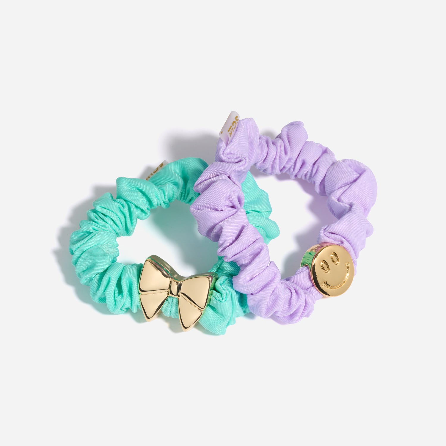 Two scrunchies, one green with a gold bow charm and one purple with a gold smiley face charm, on a white background.